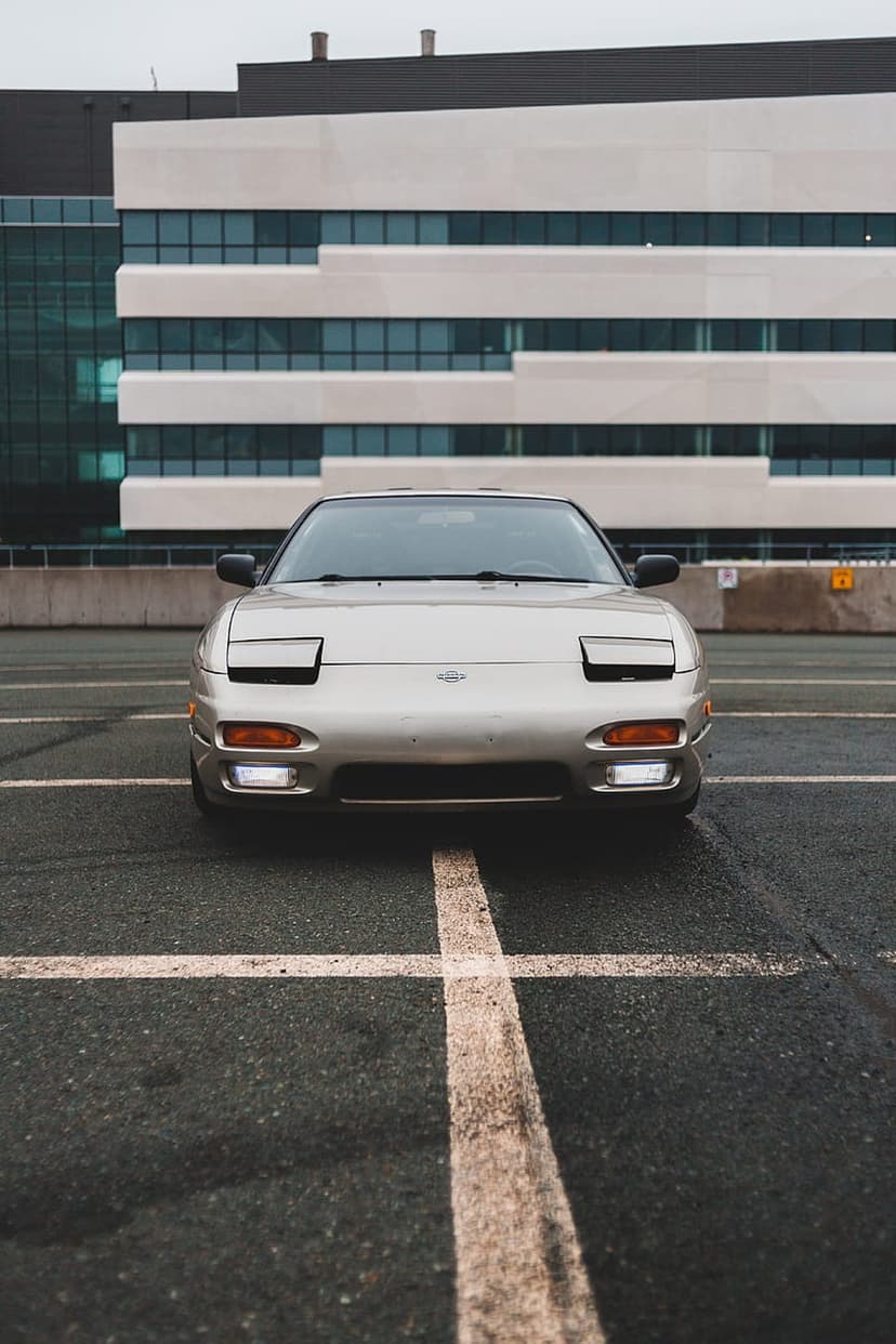 Silver Nissan 240SX S13 with pop-up headlights parked on asphalt against a modern building and overcast sky.