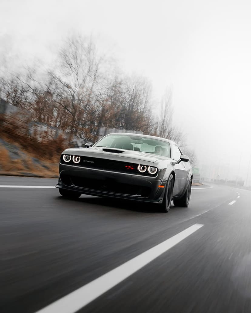Black Dodge Challenger driving on a misty road with glowing headlights and blurred trees in the background.