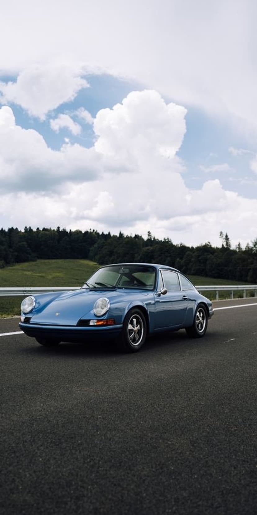 Blue vintage Porsche 911 coupe driving on a paved road between green fields and a dense forest under clouds.