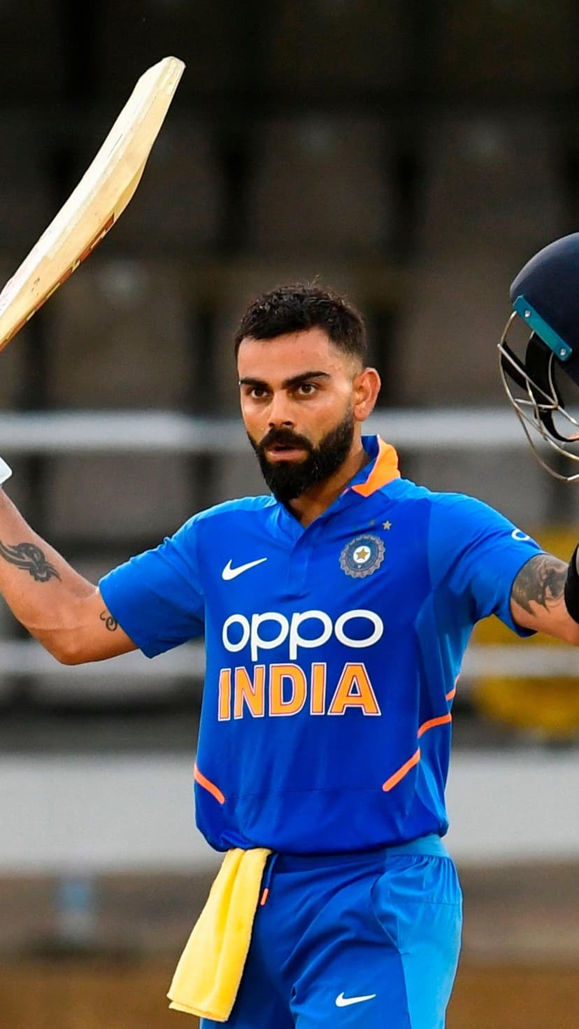 Virat Kohli raises a cricket bat and helmet in triumph wearing a blue India jersey against a blurred stadium.