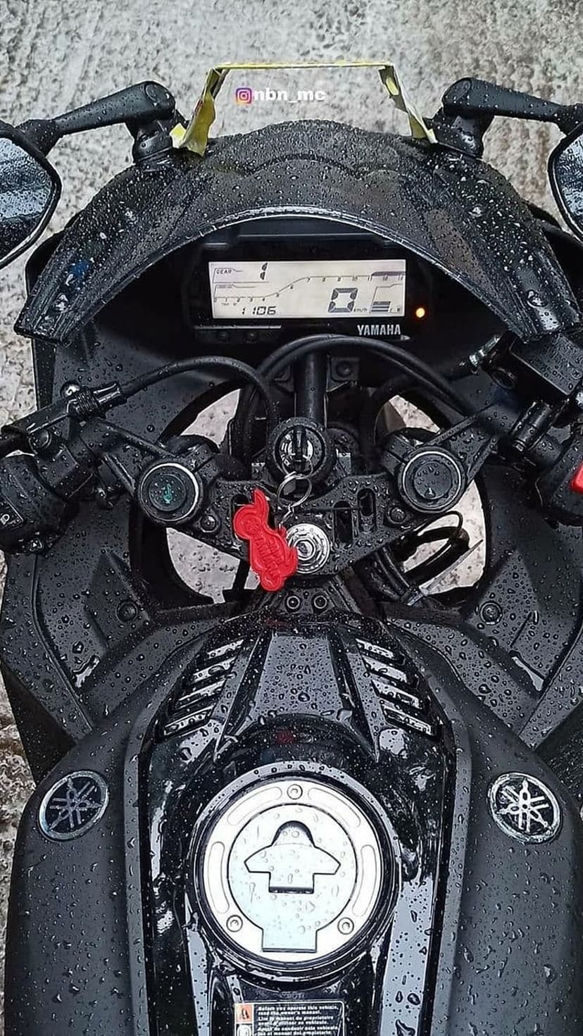 Yamaha R15 V3 handlebars and fuel tank covered in raindrops with a red cat keychain and digital speedometer.