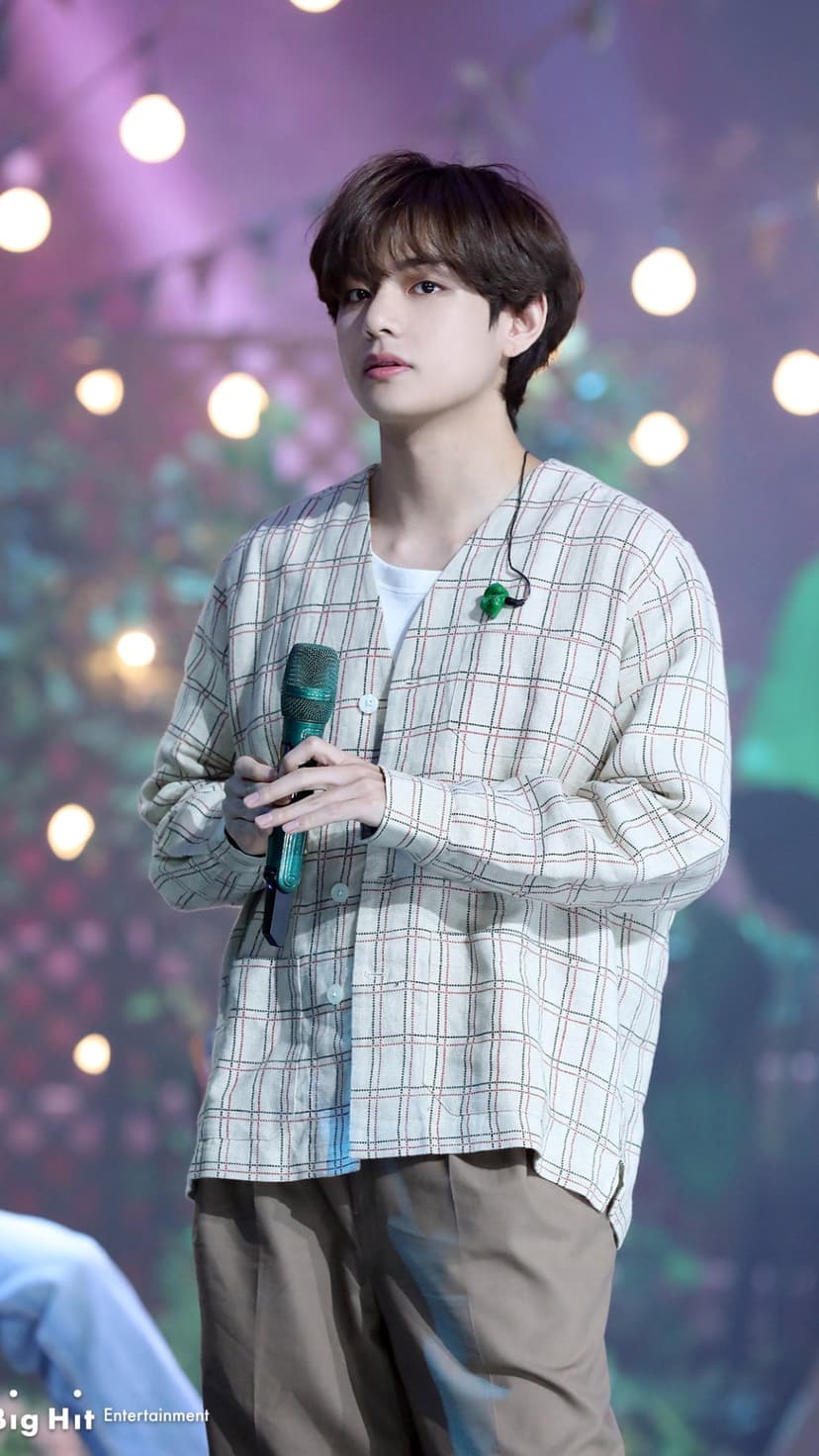 Kim Taehyung of BTS sings into a microphone wearing a checkered shirt against a soft bokeh light background.