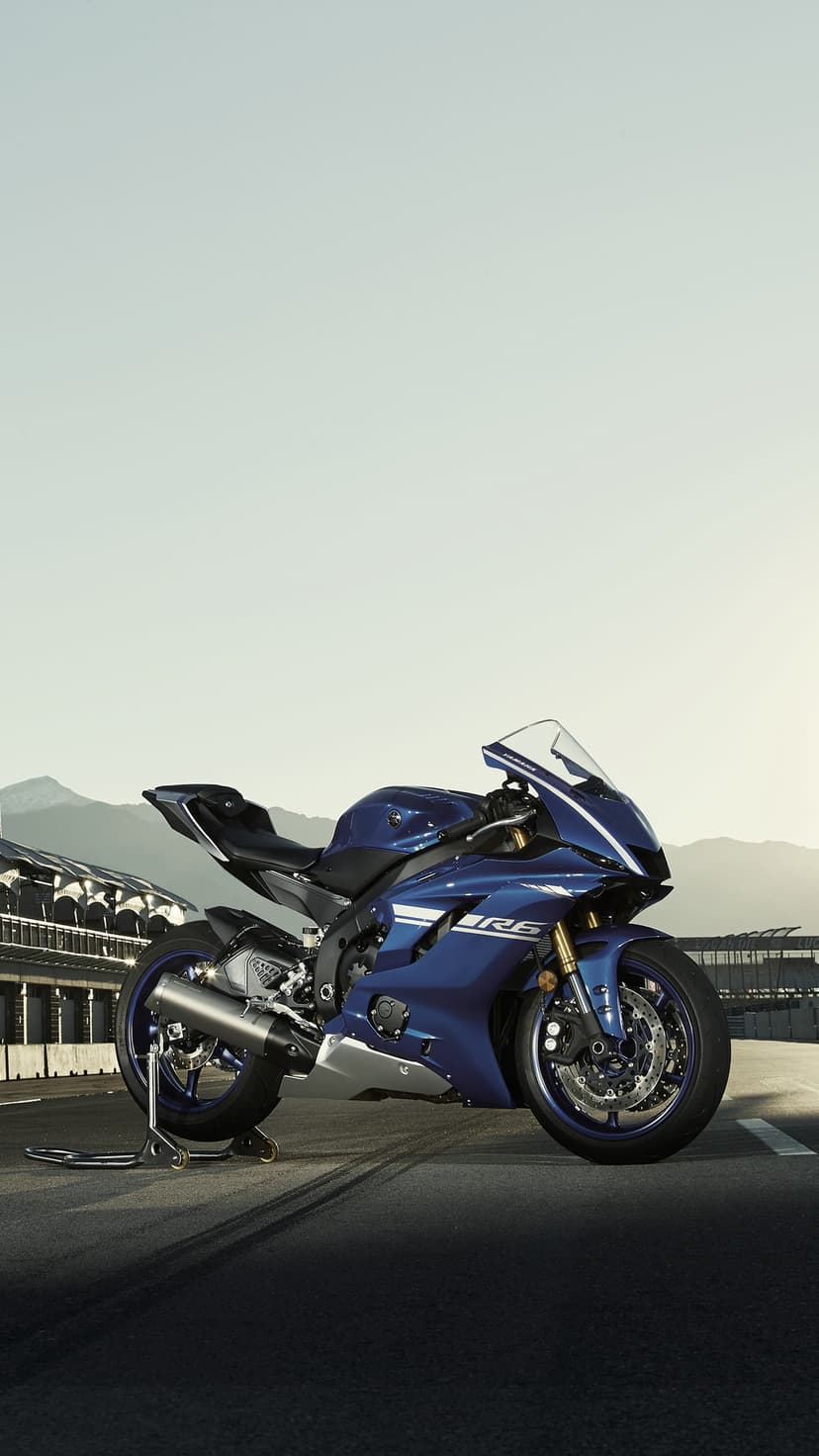 Blue Yamaha R6 motorcycle parked on a paved race track with jagged mountain peaks under a clear blue sky.