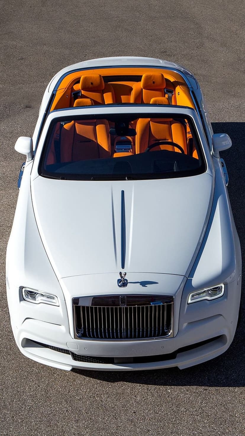White Rolls-Royce convertible with vibrant orange leather seats viewed from an aerial angle on dark asphalt.