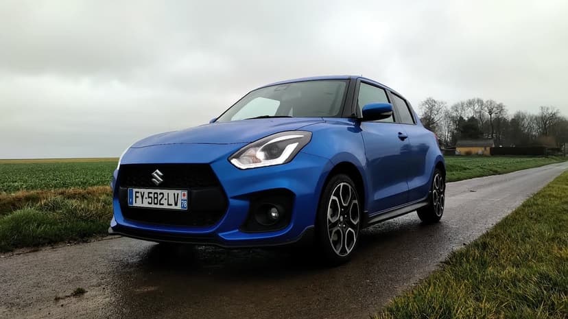 Blue Suzuki Swift Sport parked on a wet asphalt road between green fields under a cloudy overcast sky.