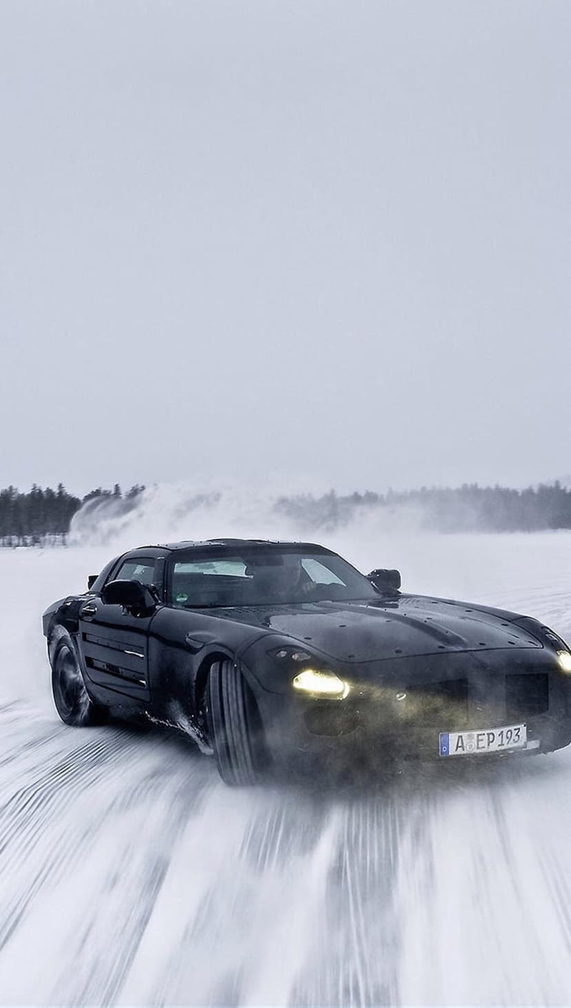 Mercedes SLS AMG drifts on a snow-covered road with headlights glowing through a hazy winter atmosphere.