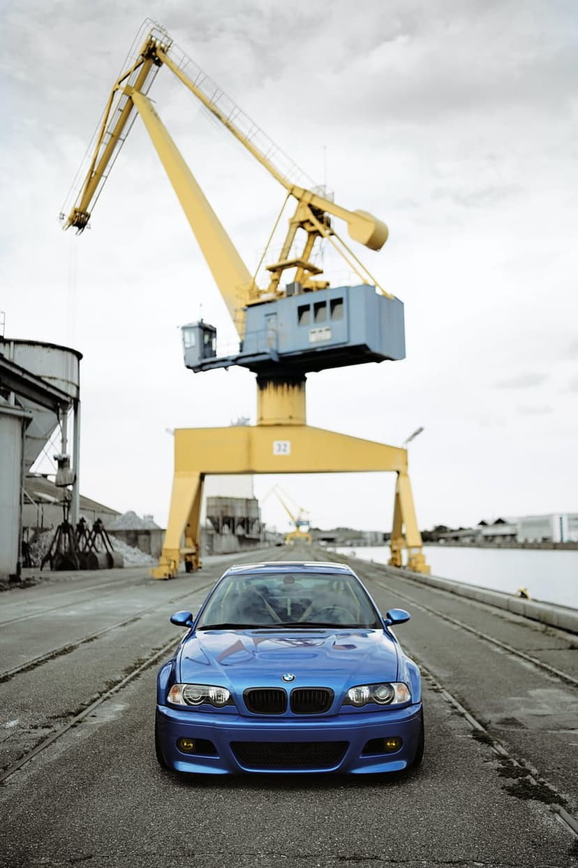 Blue BMW M3 sports car parked on asphalt at a shipping port under a large yellow gantry crane and cloudy sky.