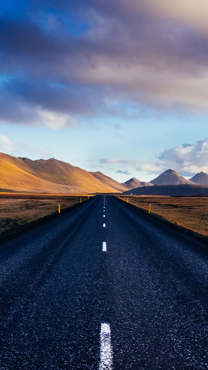 Long straight road with white markings stretching toward golden mountains under a cloudy sky in rural Iceland.
