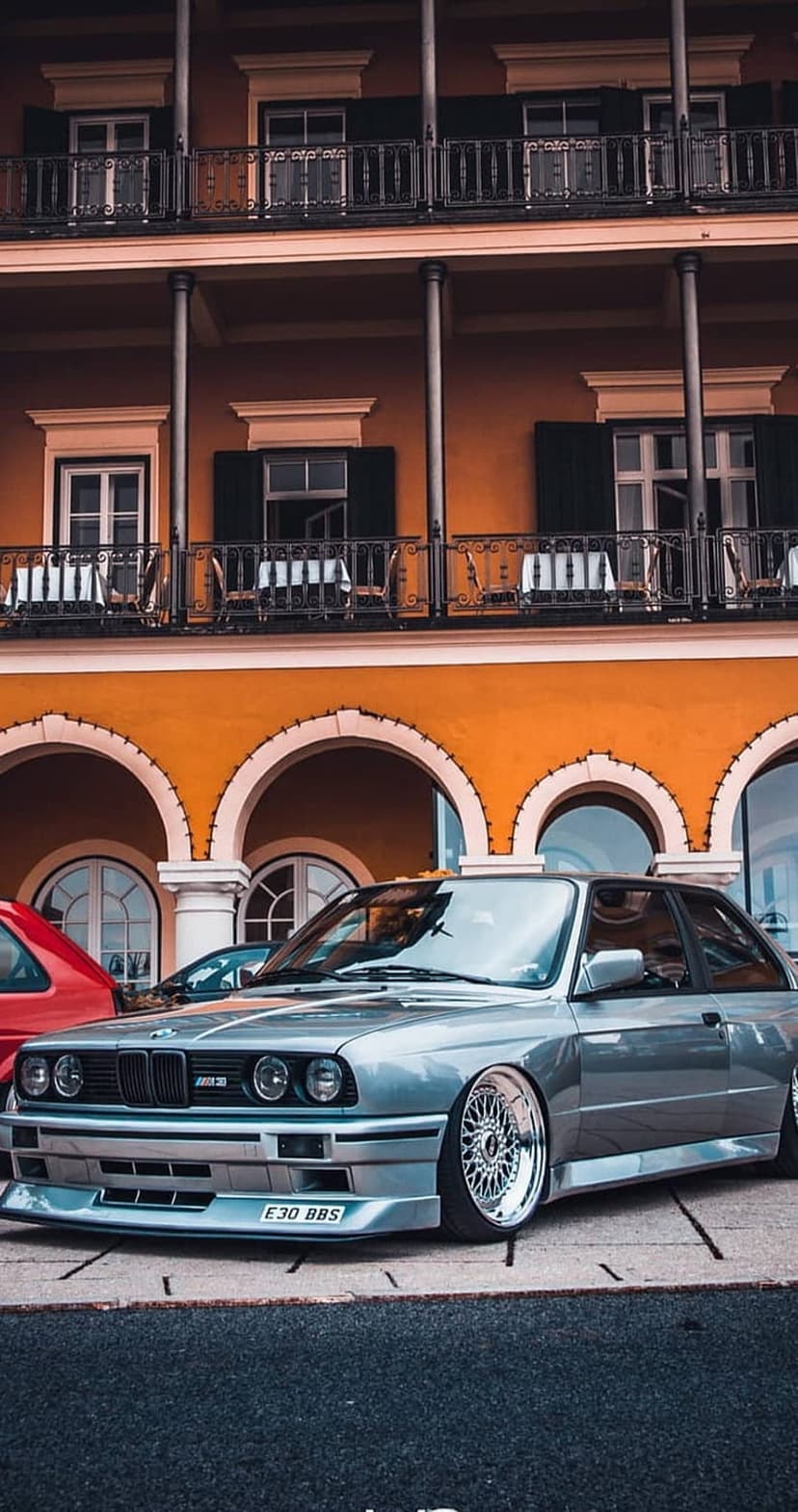 Silver BMW E30 M3 with BBS wheels parked in front of a historic stone building featuring arched window balconies.