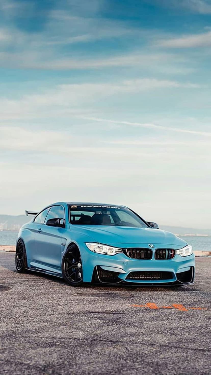 Blue BMW M4 convertible with black wheels parked on a coastal road overlooking the ocean under a cloudy sky.