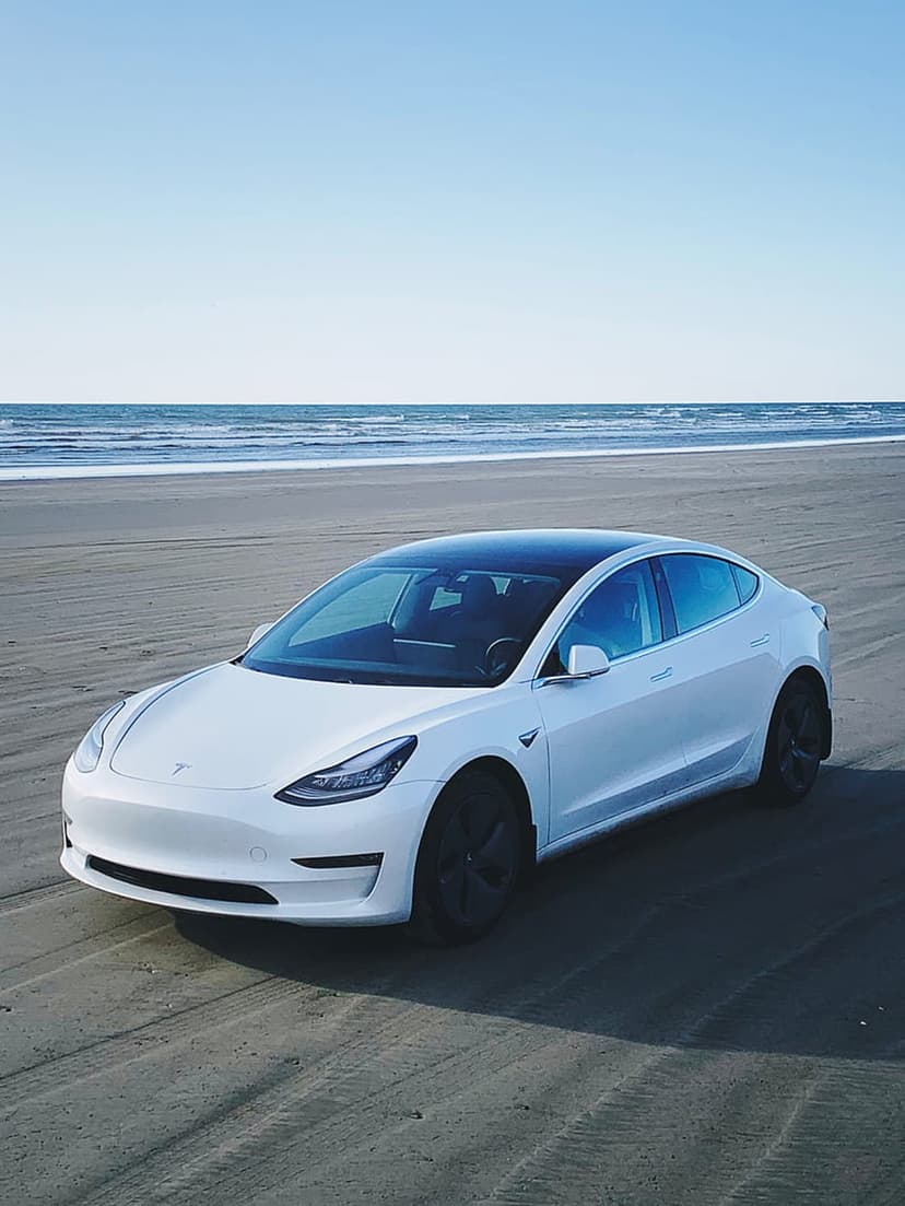 Tesla Model 3 in pearl white parked on a sandy beach with calm ocean waves under a clear blue afternoon sky.