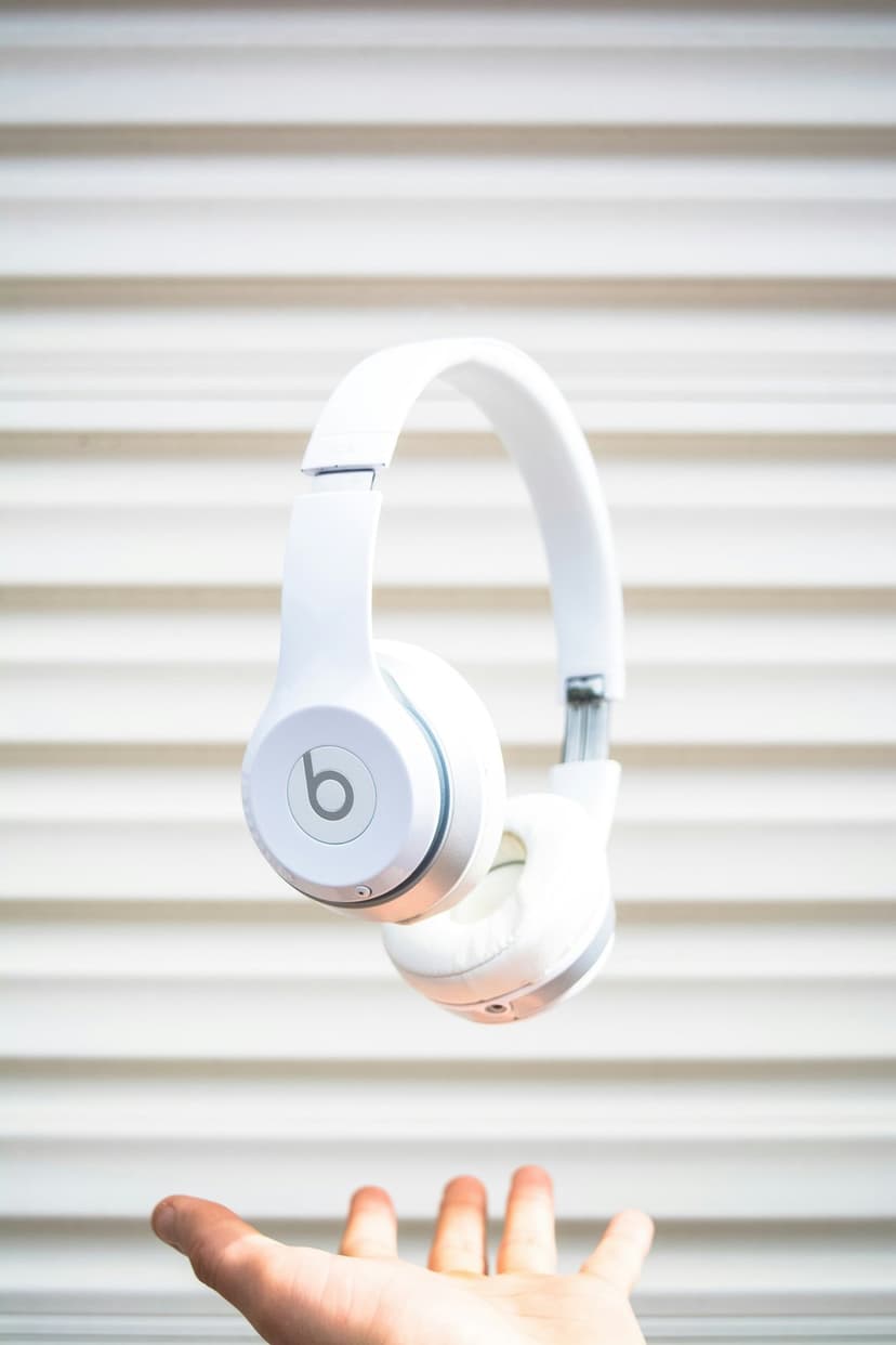 White wireless headphones floating above a human hand against a backdrop of horizontal window blinds in soft light.