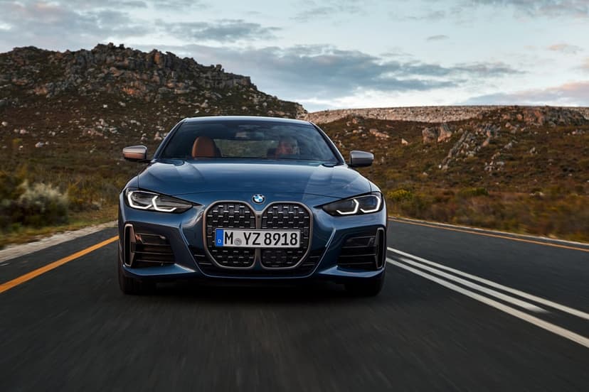 Blue BMW 4 Series coupe driving on a winding asphalt road through rocky hills during a golden sunset.