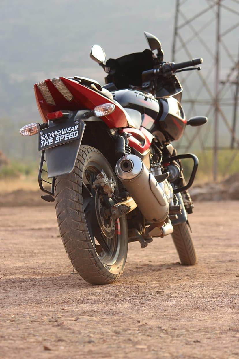 Bajaj Pulsar 220F motorcycle rear view on a dusty path with red taillights and a rugged exhaust pipe.