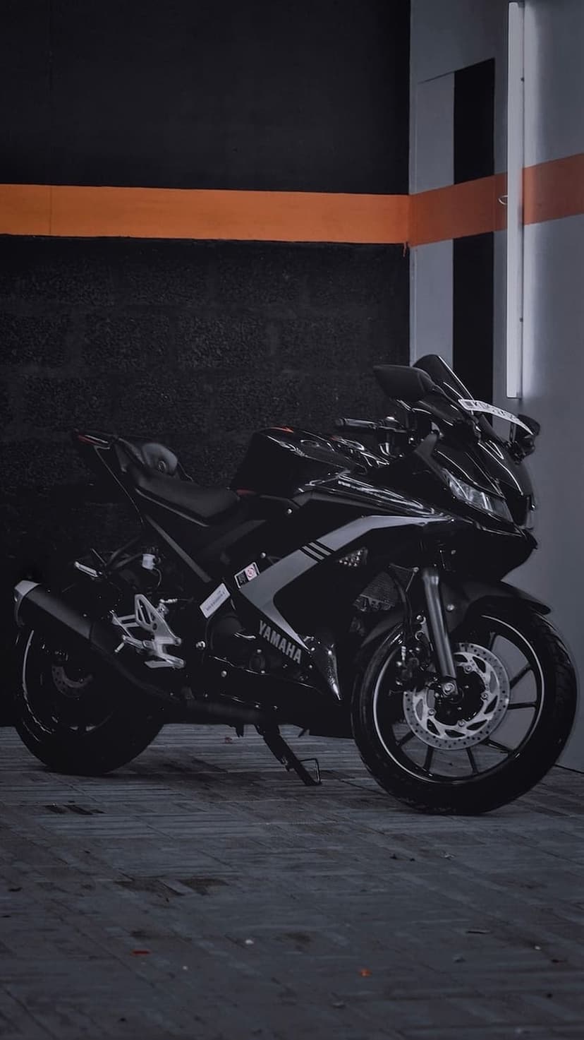 Yamaha R15 motorcycle in matte black parked next to a dark textured wall with a bright orange stripe and grey column.