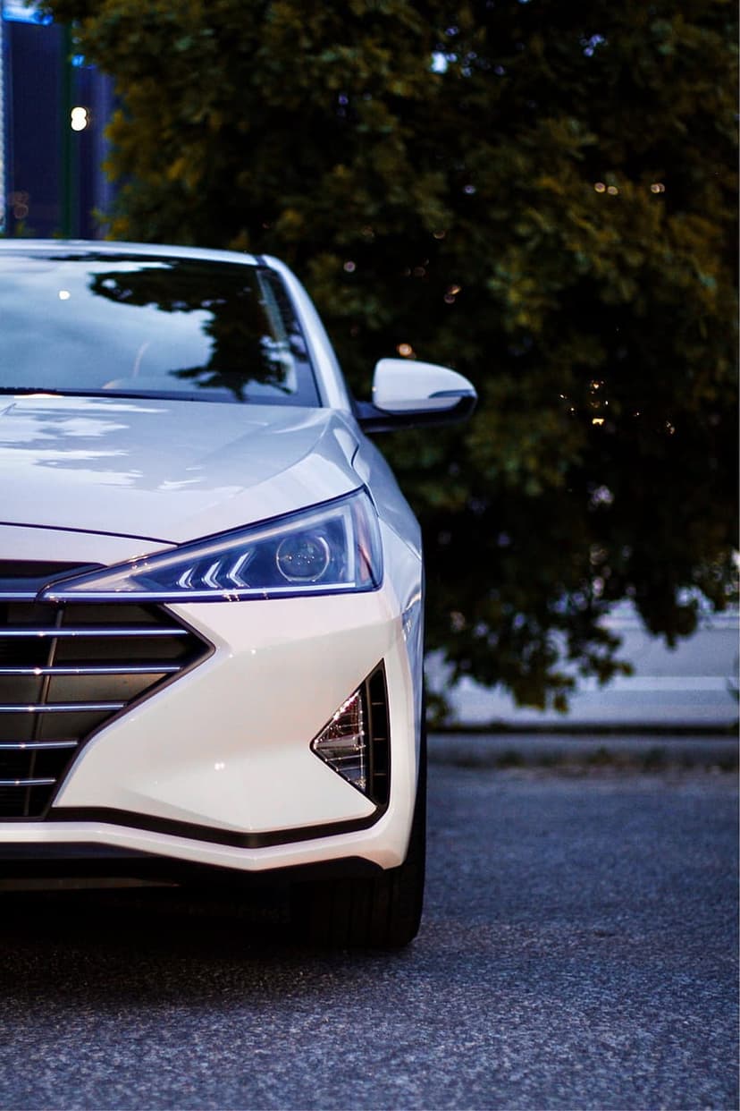 White Hyundai Elantra headlight and chrome grille shown in a low-angle close-up against a soft city bokeh at dusk.
