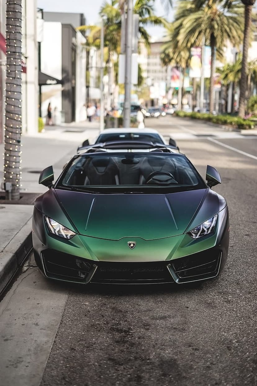 Green iridescent Lamborghini convertible parked on a sunlit street with palm trees and shops in the background.