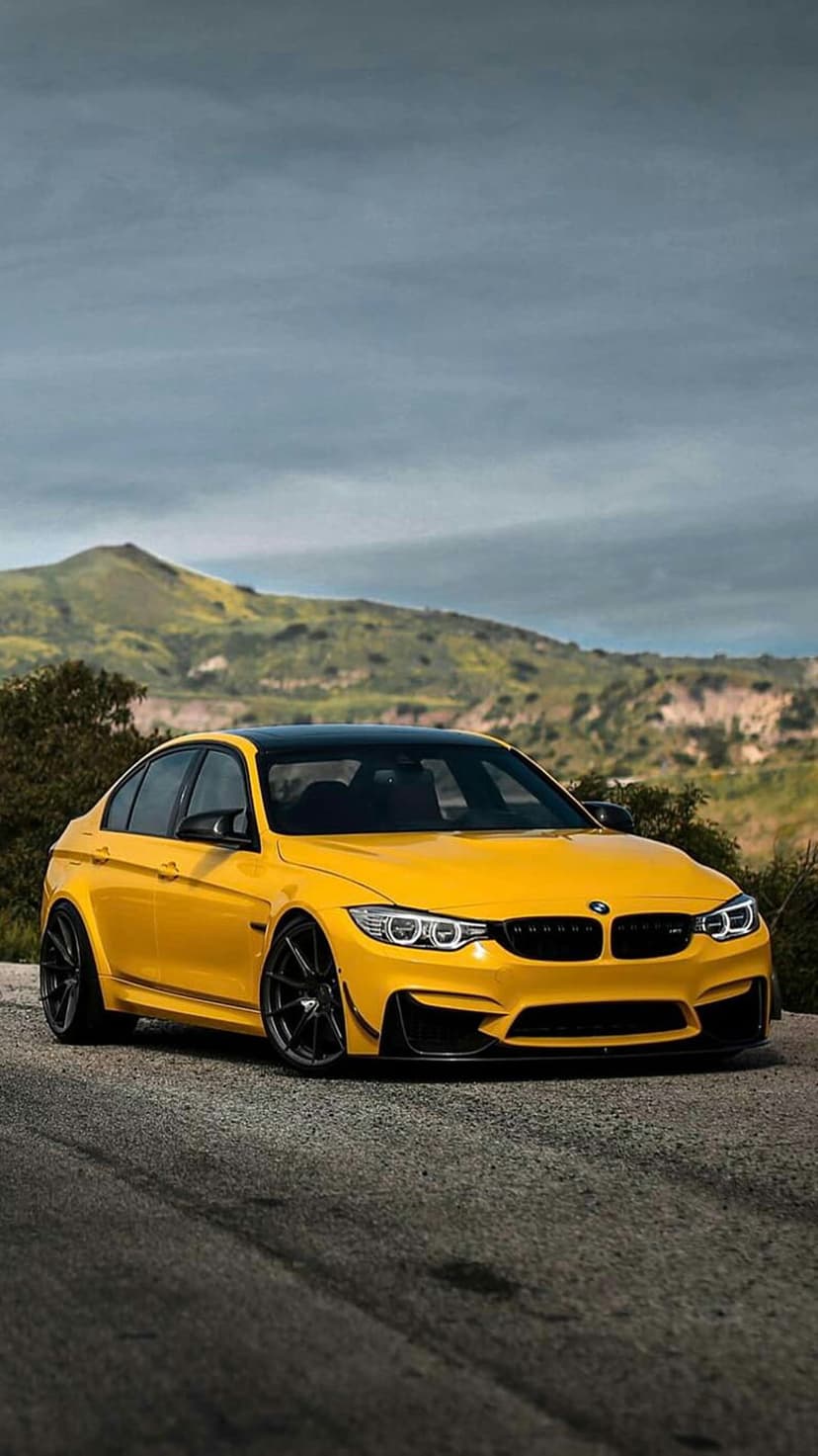 Yellow BMW M4 Coupe on an asphalt road with green mountains and a cloudy sky in the background under daylight.
