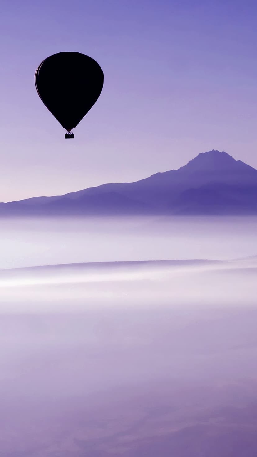 Hot air balloon silhouette drifting over a sea of purple clouds with a distant mountain peak on the horizon.