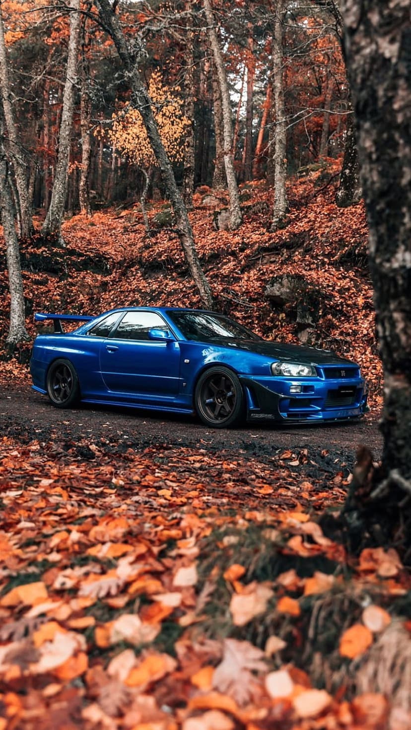 Blue Nissan Skyline R34 parked on a dirt road covered in orange autumn leaves within a misty forest setting.