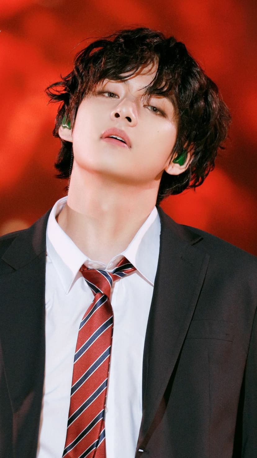 Kim Taehyung wears a black suit, striped tie, and green earbuds against a blurred red background in this portrait.