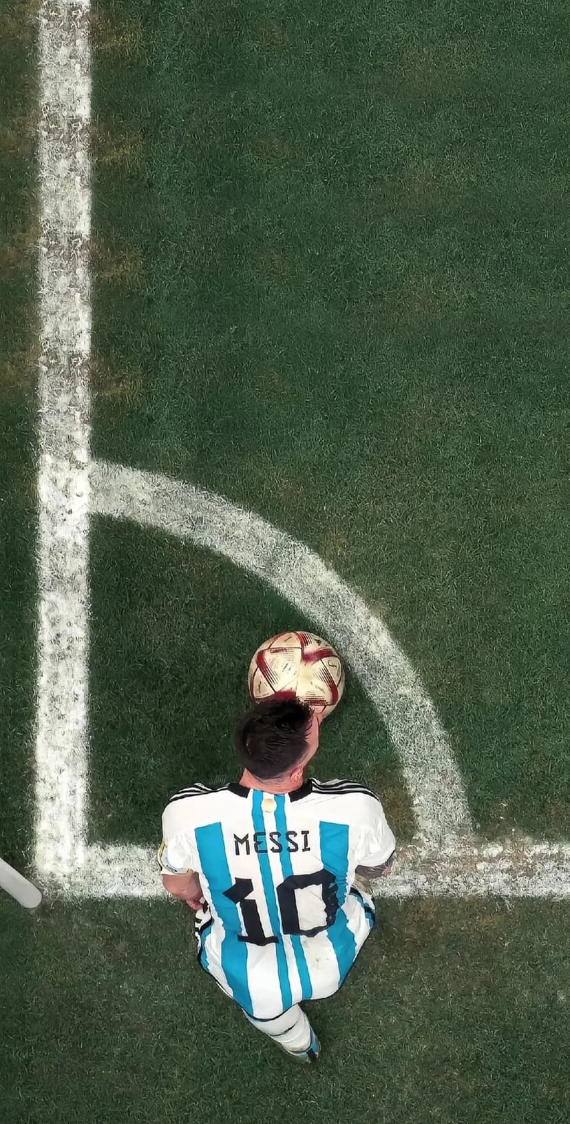 Lionel Messi in his striped Argentina kit sits on a grass field near a soccer ball and white corner line.