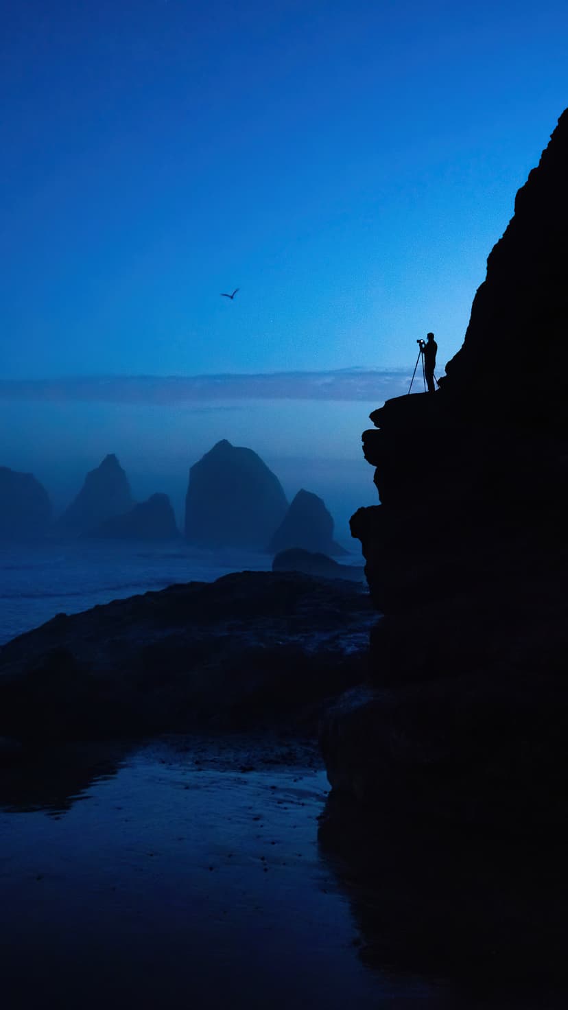 Silhouette of a photographer on a steep cliff overlooking misty sea stacks and a dark blue ocean at twilight.