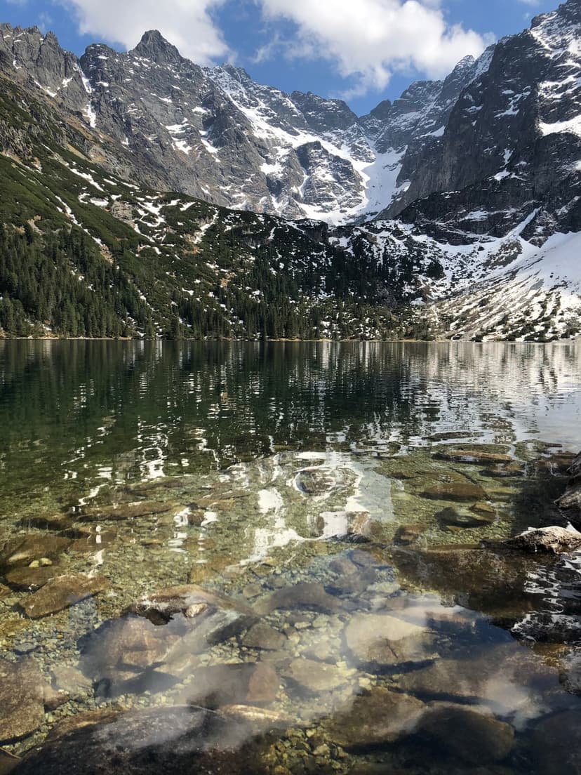 Snow-capped mountain peaks towering over a calm lake with evergreen trees and a bright blue sky reflected in water.