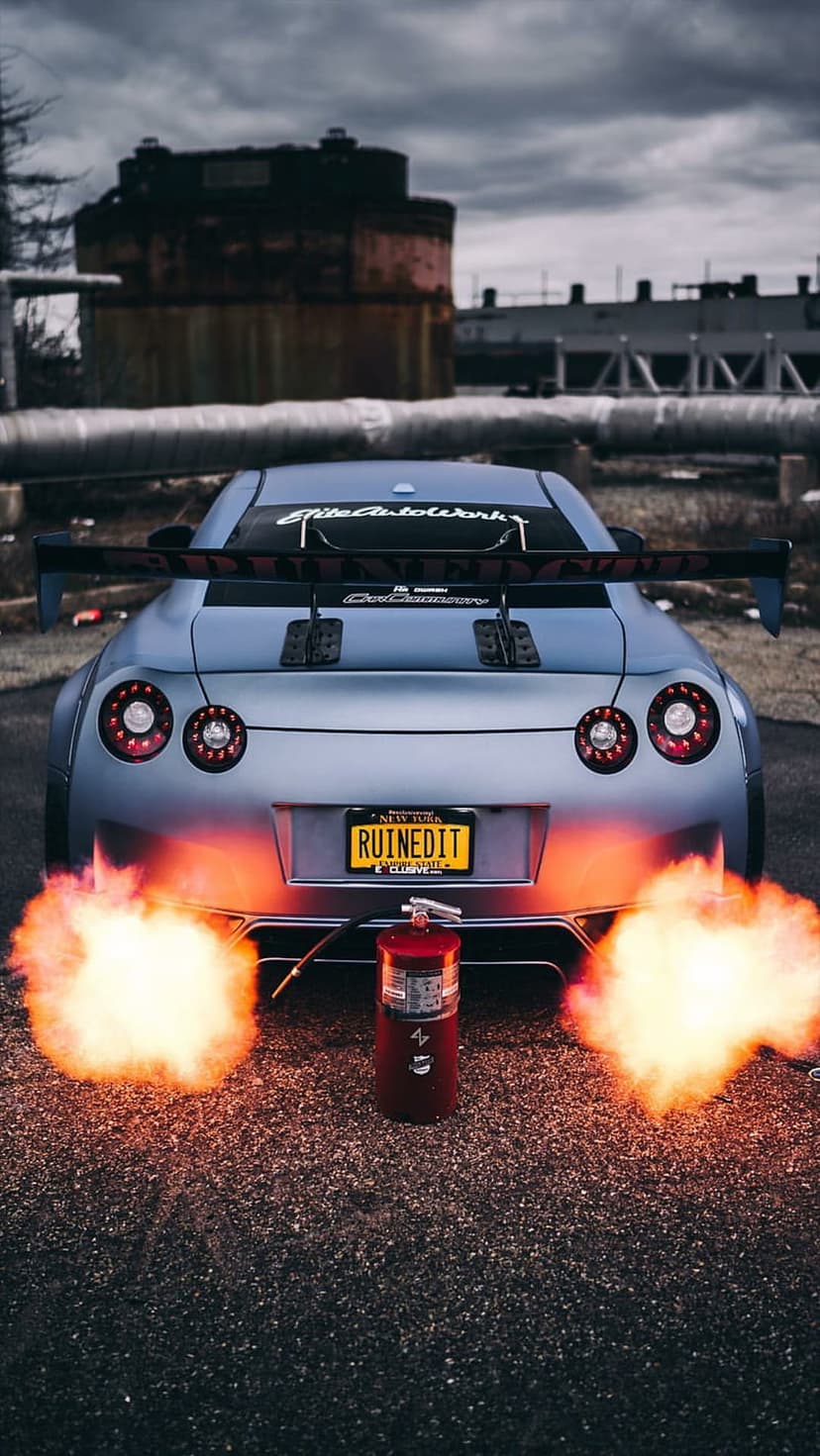 Nissan GT-R with a Ruined It plate shoots orange exhaust flames near a red fire extinguisher on asphalt.