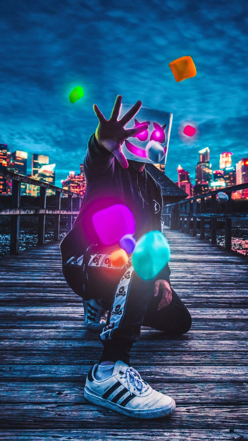 Marshmello DJ dancing with colorful glowing orbs on a wooden pier with a bright city skyline backdrop at night.