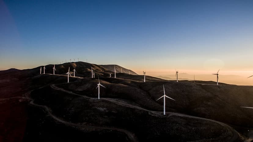 Wind turbine silhouettes stand on dark rolling hills against a vibrant orange and purple sunset sky at twilight.