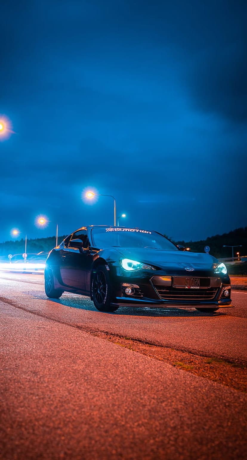 Subaru BRZ drifting on wet asphalt at night with glowing blue headlights and orange streetlamp reflections.