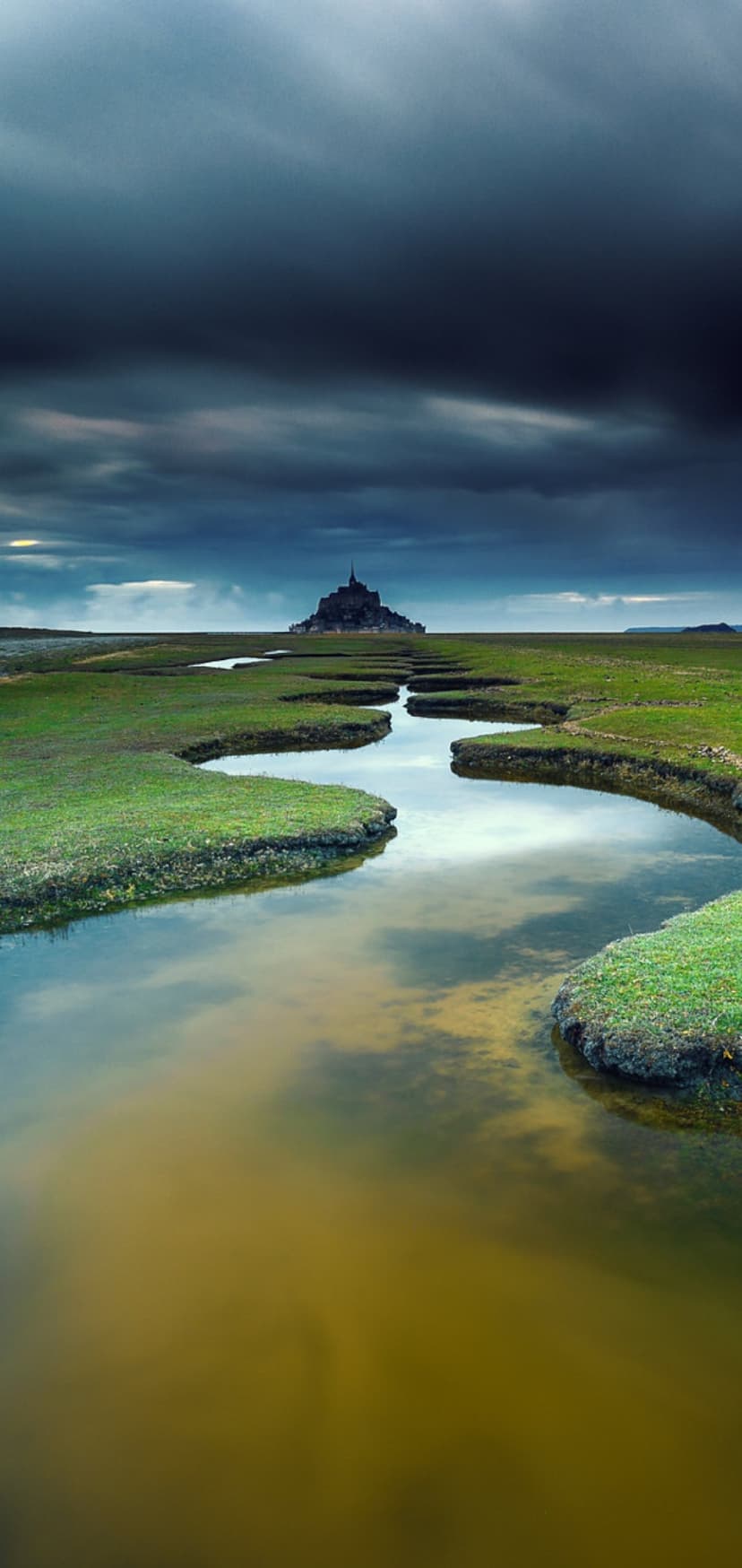 Mont Saint-Michel abbey fortress stands amidst green fields and winding rivers under heavy storm clouds.