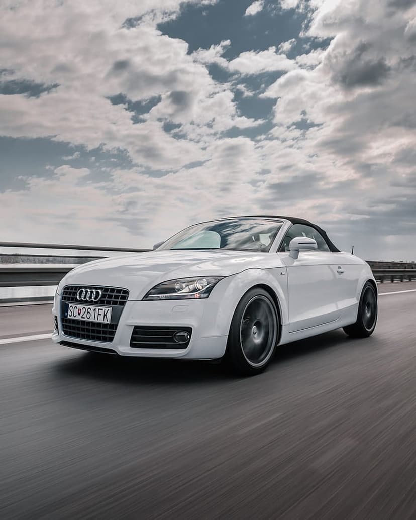 White Audi TT convertible sports car driving on a highway with motion blur beneath a dramatic cloudy sky.