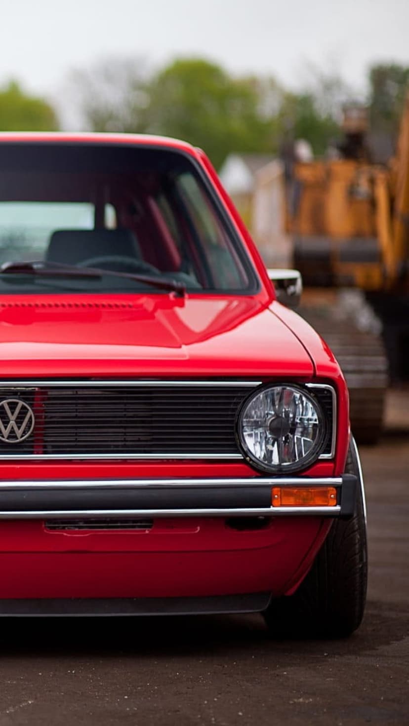 Red Volkswagen Golf MK1 front end featuring a round headlight and VW badge against a blurred green background.