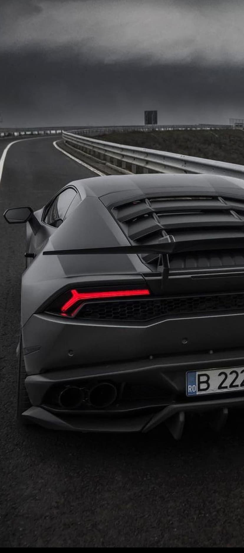 Black Lamborghini rear with glowing red taillights on a dark winding road under a cloudy stormy sky at dusk.