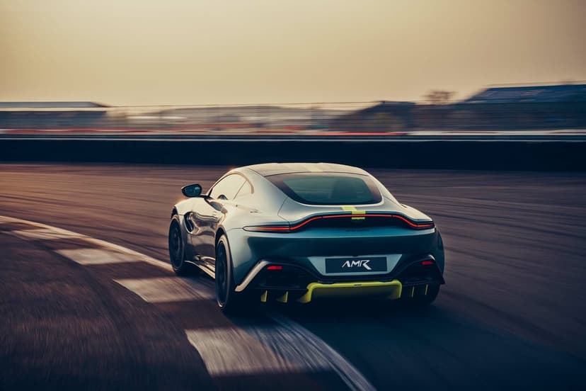 Silver Aston Martin Vantage AMR with lime yellow accents speeding on a racetrack under warm sunset lighting.