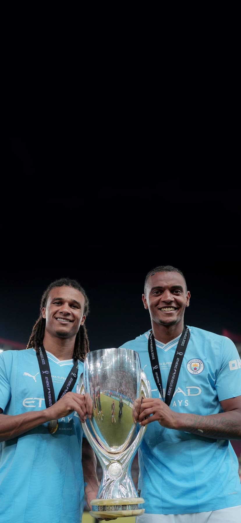 Manchester City players in blue kits hold a silver trophy while wearing gold medals against a dark background.