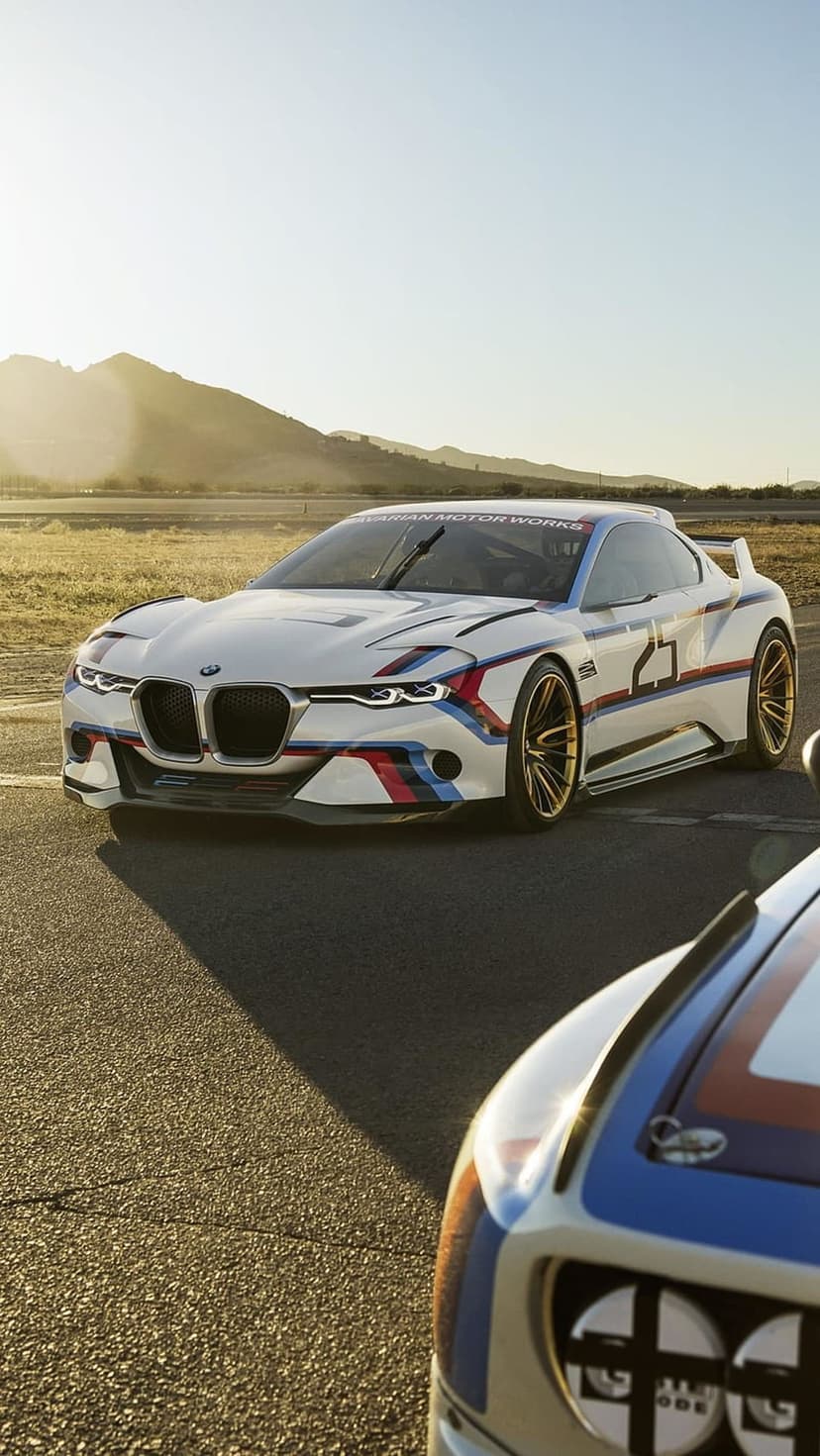 BMW 3.0 CSL Hommage R race car with number 25 and gold wheels on an asphalt track against a mountain backdrop.