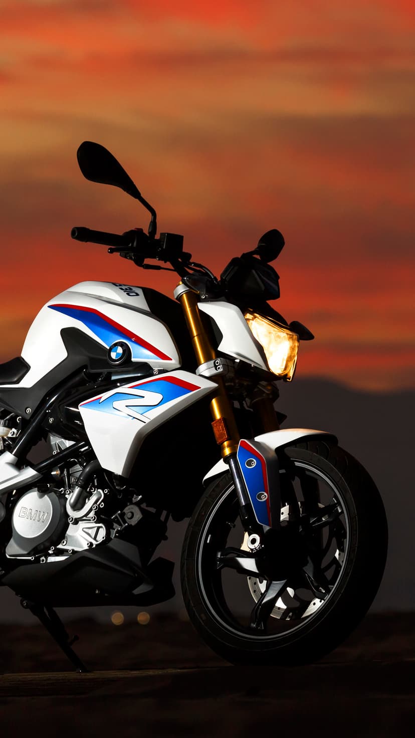White BMW motorcycle with glowing headlight parked against a purple mountain sunset during twilight hours.