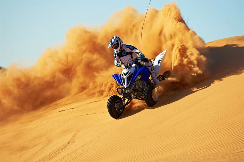Blue quad bike rider drifting on a golden sand dune, creating a massive orange dust cloud under a clear sky.