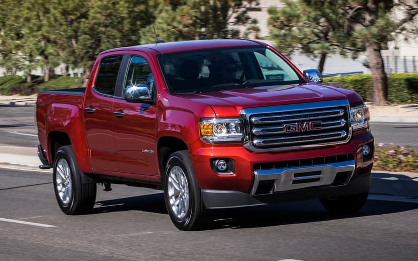 Red GMC Canyon pickup truck driving on an asphalt road with blurred green trees and buildings behind it.