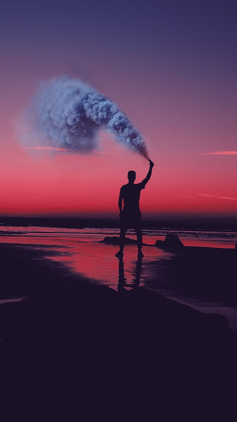 Silhouetted person holds a white smoke flare on a wet beach during a vibrant pink and purple sunset.