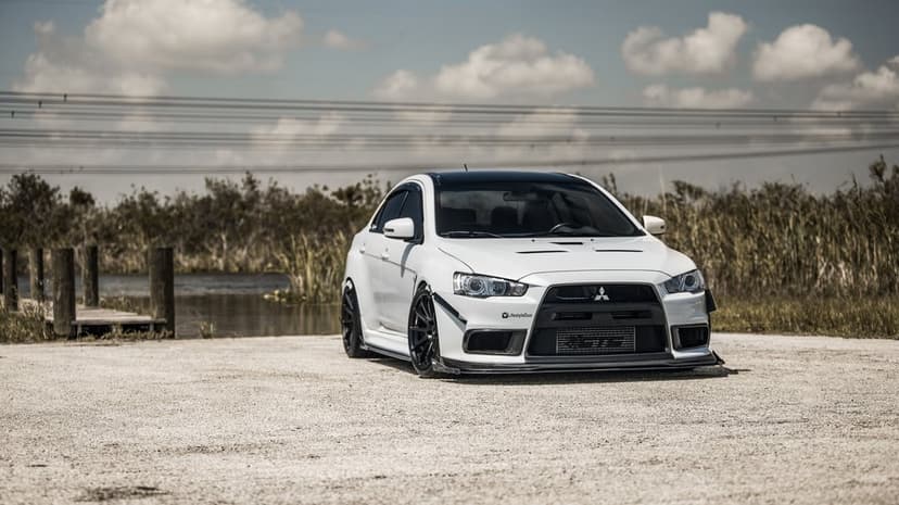 White modified Mitsubishi Lancer Evo X parked on a light gravel path near a lake under a desaturated sky.