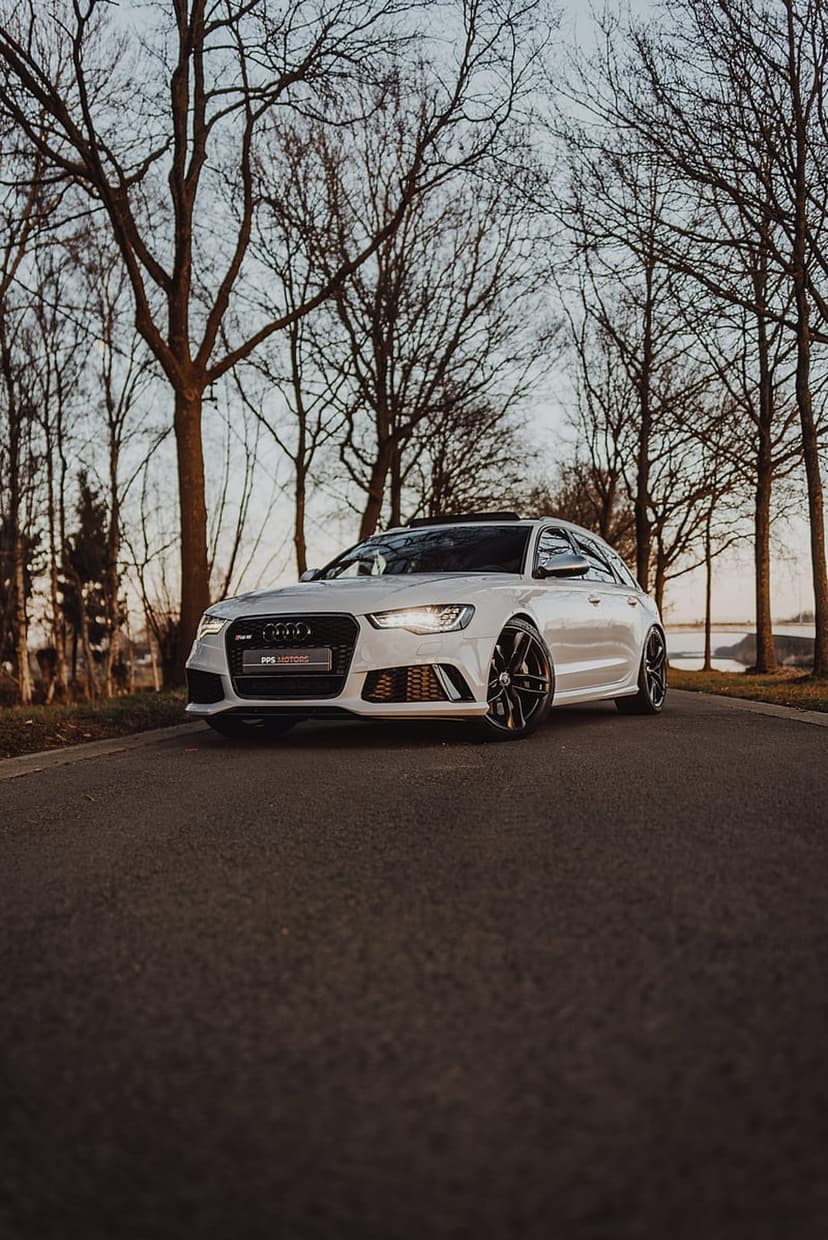 White Audi RS6 Avant with black wheels parked on a paved forest road under a hazy twilight sky during winter.