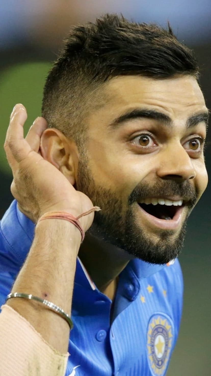 Virat Kohli in a blue India jersey cups his hand to his ear while cheering during a professional cricket match.