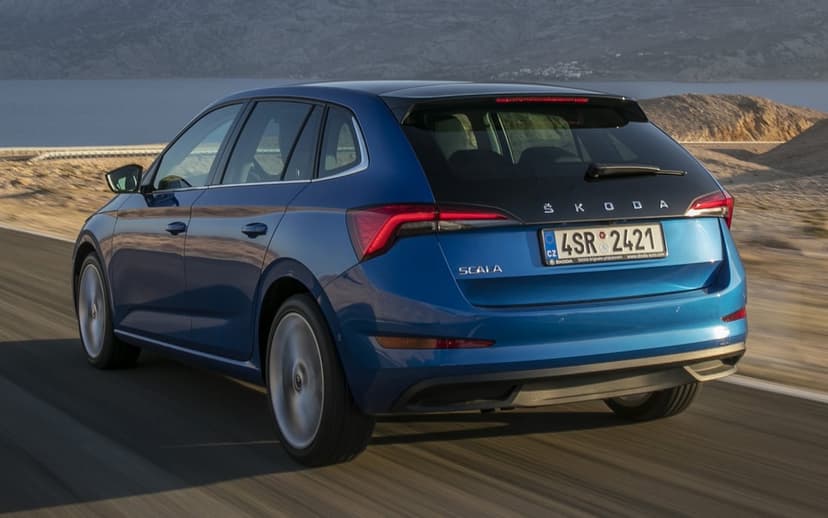 Blue Skoda Scala hatchback driving on a scenic coastal road with mountains and water in the background at sunset.