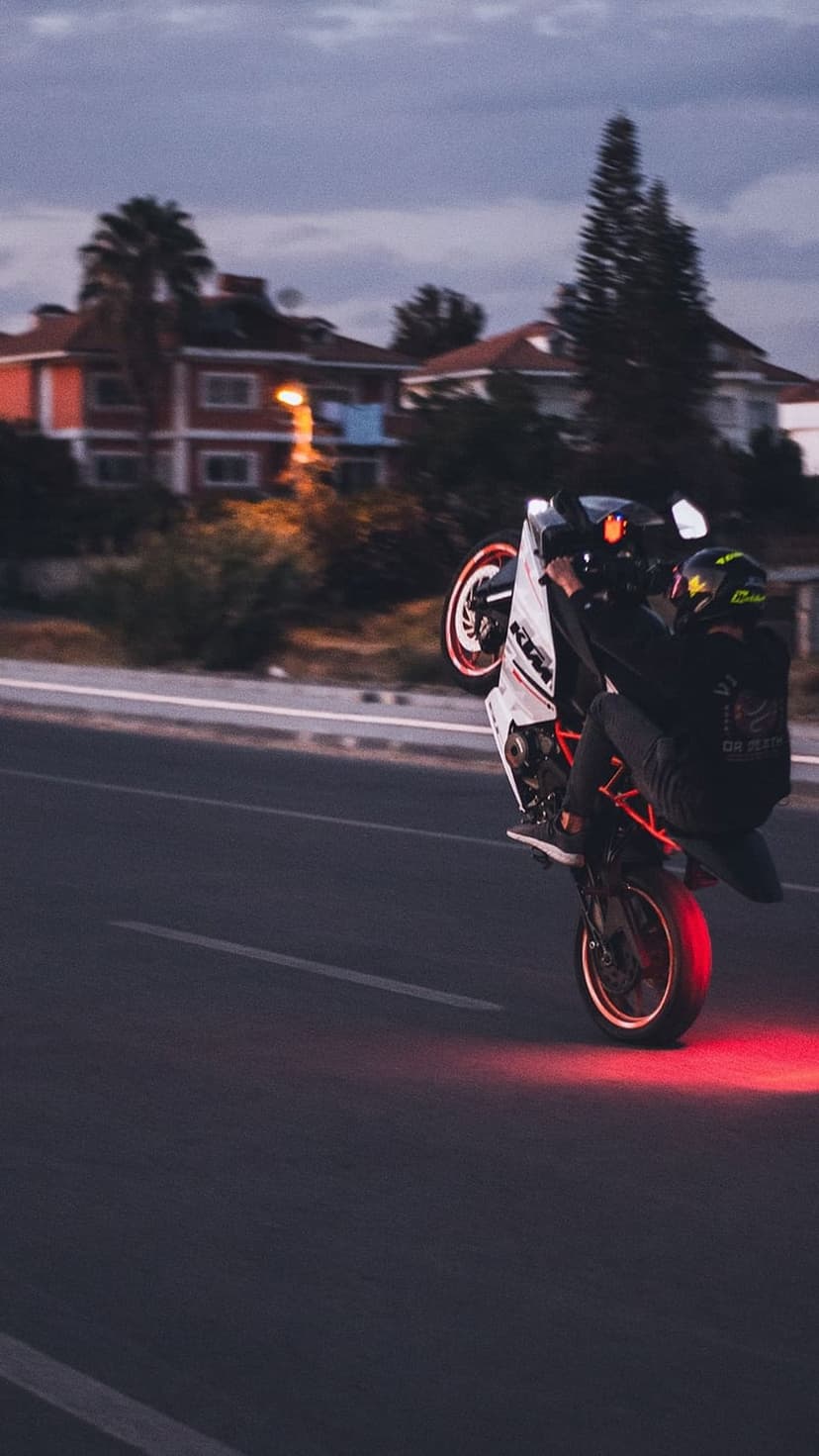 Motorcyclist in a wheelie on a dark street as red tail lights cast a vibrant glow on the asphalt at dusk.