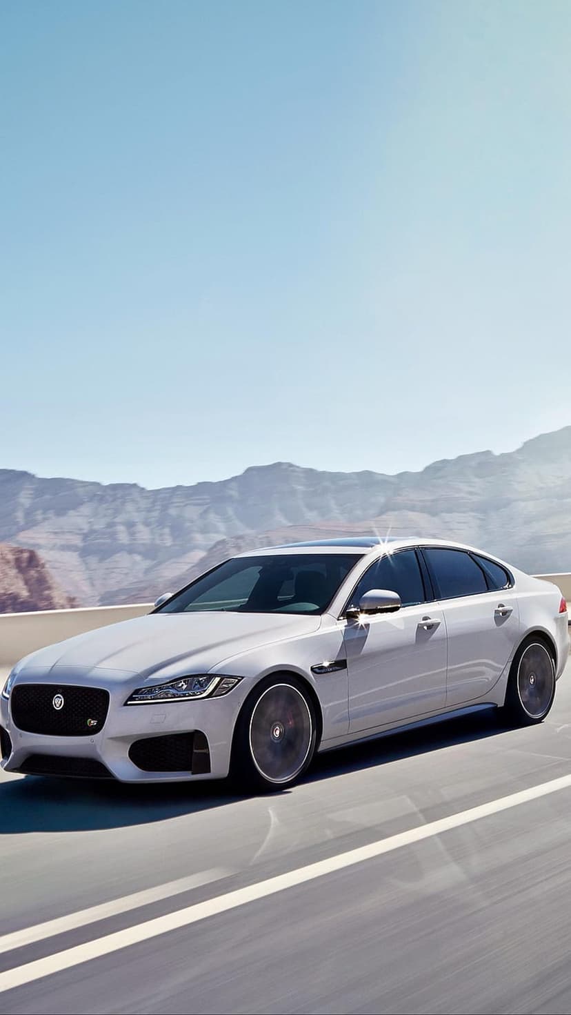 White Jaguar XF sedan driving on a paved mountain road against a backdrop of rocky peaks and a clear blue sky.