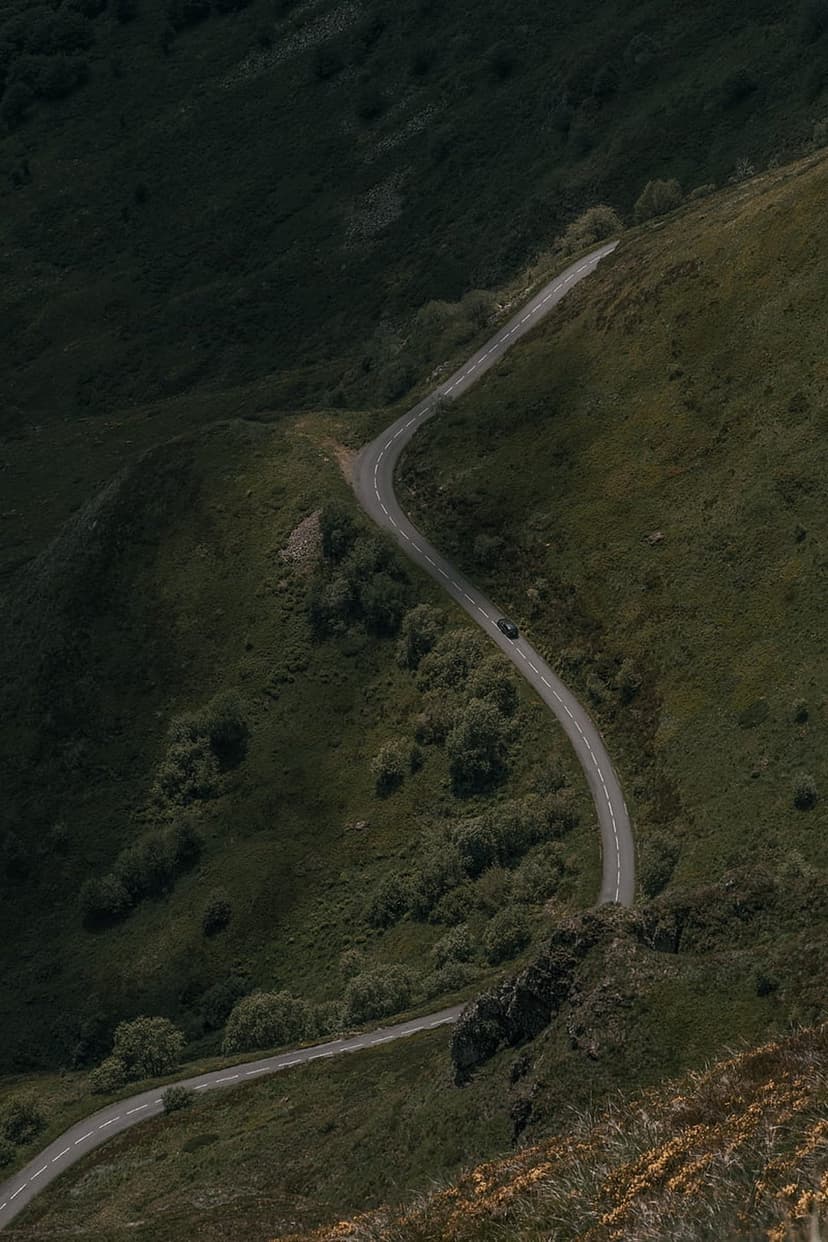 Winding asphalt road curves through steep green hills with a single white car driving along the mountain ridge.